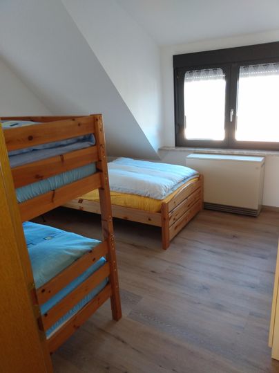 Ein einfaches Schlafzimmer mit einem Etagenbett und einem Einzelbett. Es hat ein Fenster, das viel Tageslicht hereinlässt.