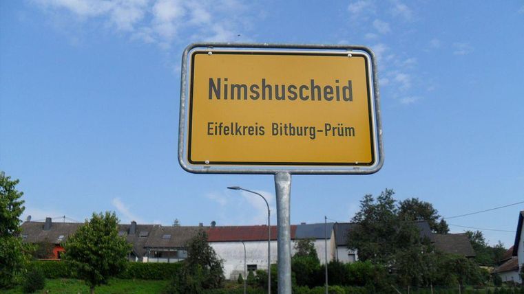 Een plaatsnaambord van Nimshuscheid in de Eifelkreis Bitburg-Prüm. De omgeving is landelijk met groene weilanden en huizen op de achtergrond.
