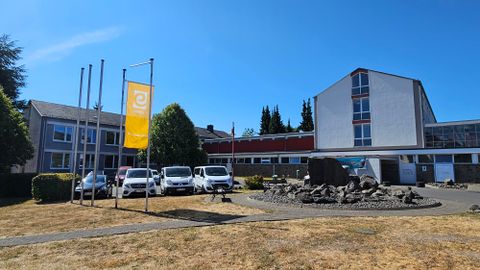 Ein modern gestaltetes Gebäude mit großen Fenstern und einem Glockenturm. Im Vordergrund ist ein begrüntes Gelände mit Bäumen und Straßenlaternen zu sehen.