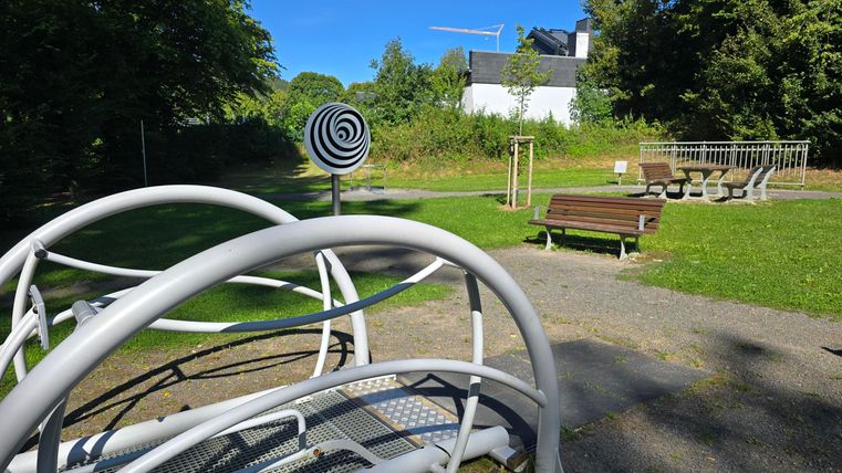 Ein Spielplatz mit verschiedenen Geräten und einer grünen Wiese. Im Hintergrund sind Bäume und ein Gebäude zu sehen.
