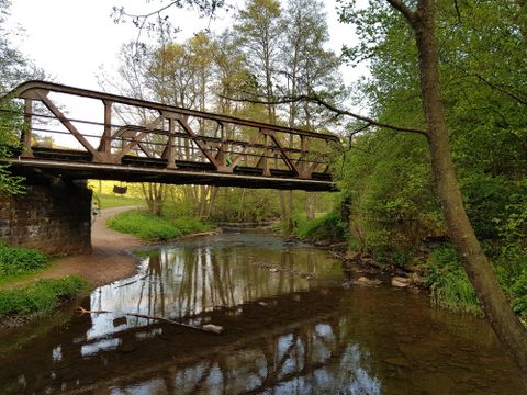 An old wooden bridge over a small river. Green trees and bushes surround the scene.