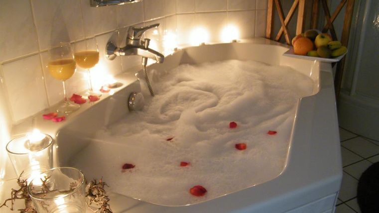 Eine entspannende Badewanne mit Schaum und Rosenblättern. Kerzen und Obst schaffen eine gemütliche Atmosphäre.