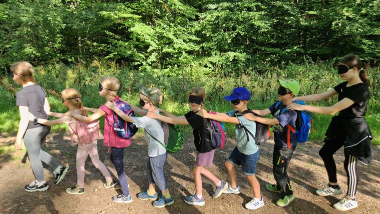 Eine Gruppe von Kindern geht im Wald, während sie Augenbinden tragen. Sie sind mit Rucksäcken ausgestattet und halten sich an den Schultern fest.