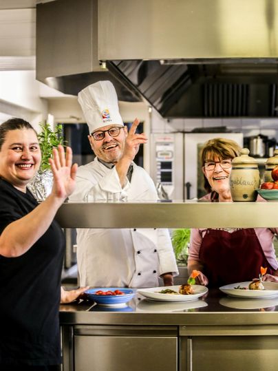 Drei Köche lächeln und winken in einer modernen Küche. Auf dem Tisch sind einige Teller mit zubereiteten Gerichten zu sehen.
