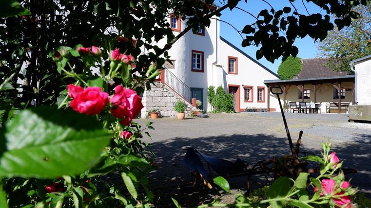 Ein idyllischer Innenhof mit blühenden Rosen und einer einladenden weißen Hausfassade. Im Hintergrund sind Gartenmöbel und ein sonniger Himmel zu sehen.