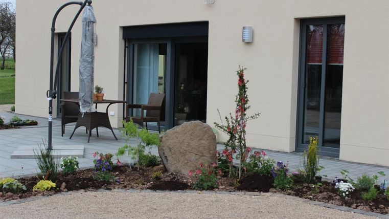 Ein schöner Eingangsbereich mit einer Terrasse, einem großen Stein und bunten Blumen. Das moderne Gebäude hat große Fenster, die viel Licht hereinlassen.