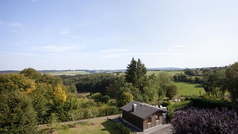 Eine malerische Landschaft mit sanften Hügeln und üppigem Grün. Im Vordergrund steht ein kleines Haus unter einem blauen Himmel.