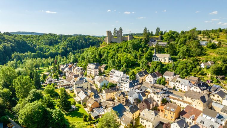 Eine malerische Stadt umgeben von grünen Wäldern. Im Hintergrund ist eine imposante Burg sichtbar.