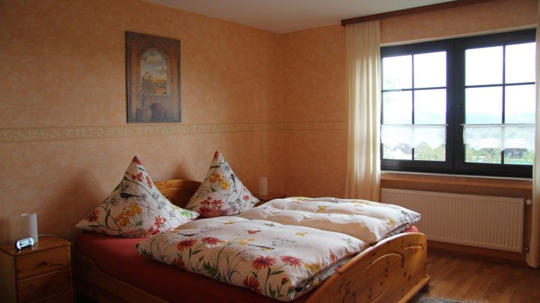 Ein gemütliches Schlafzimmer mit einem Doppelbett und bunten Kopfkissen. Große Fenster sorgen für viel Tageslicht und eine angenehme Atmosphäre.