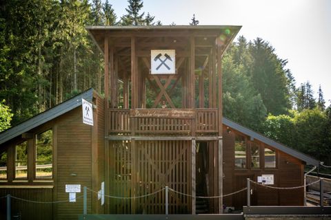 Ein historisches Bergwerkgebäude aus Holz mit einem Aussichtsturm. Umgeben von Bäumen in einer natürlichen Umgebung.