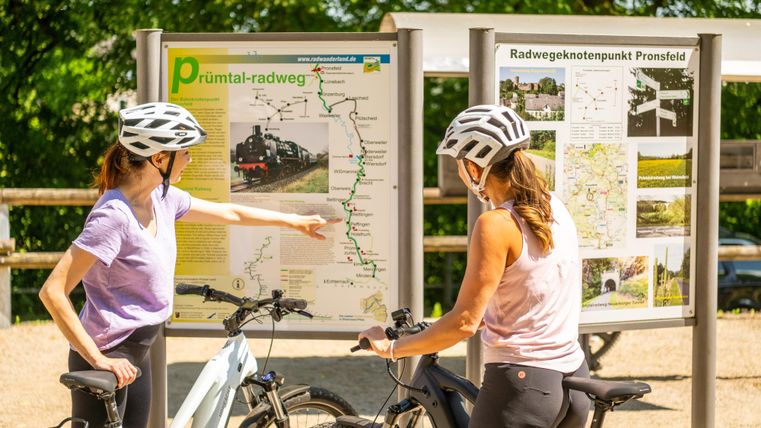 Zwei Radfahrerinnen in Helmen schauen auf eine Karte des Prüm-Tal-Radwegs. Sie planen ihre Route und scheinen sich über den Wegverlauf zu unterhalten.