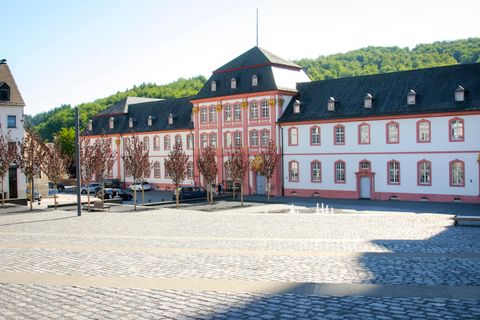 Ein historisches Gebäude mit rotem und weißem Mauerwerk steht an einem Platz. Im Hintergrund sind grüne Hügel und ein klarer Himmel zu sehen.