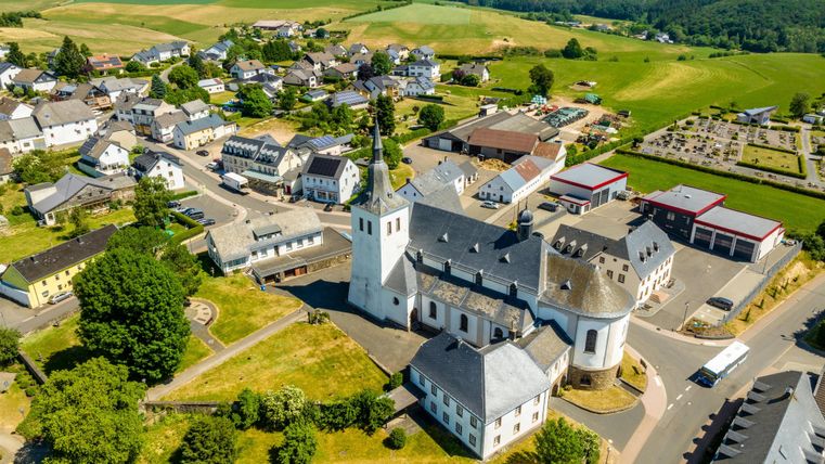 Une vue pittoresque du village avec plusieurs bâtiments et une église. À l'arrière-plan, on peut voir des champs verts et des collines douces.