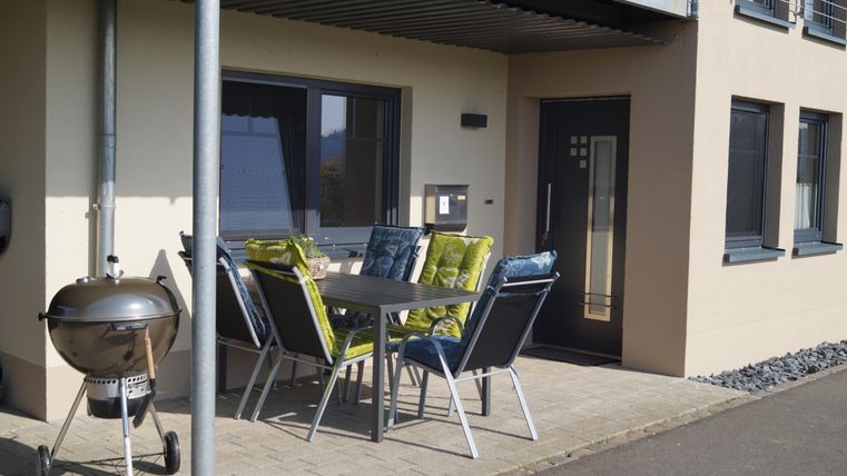 Ein moderner Außenbereich mit einem Tisch und Stühlen auf einer Terrasse. Neben dem Tisch steht ein Grill.