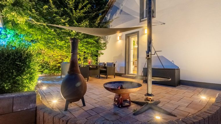 Ein gemütlicher Außenbereich mit einem Kamin und einer Feuerstelle. Die Beleuchtung und die grüne Umgebung schaffen eine einladende Atmosphäre.