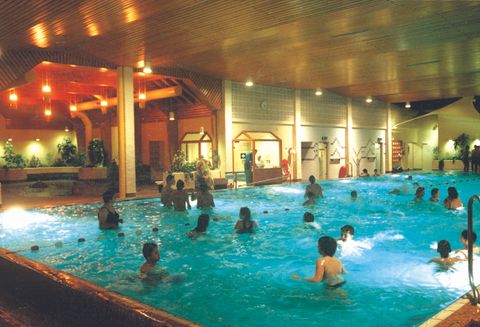 Ein Innenpool mit vielen Menschen, die im Wasser schwimmen und spielen. Die Umgebung ist hell erleuchtet und hat eine entspannte Atmosphäre.