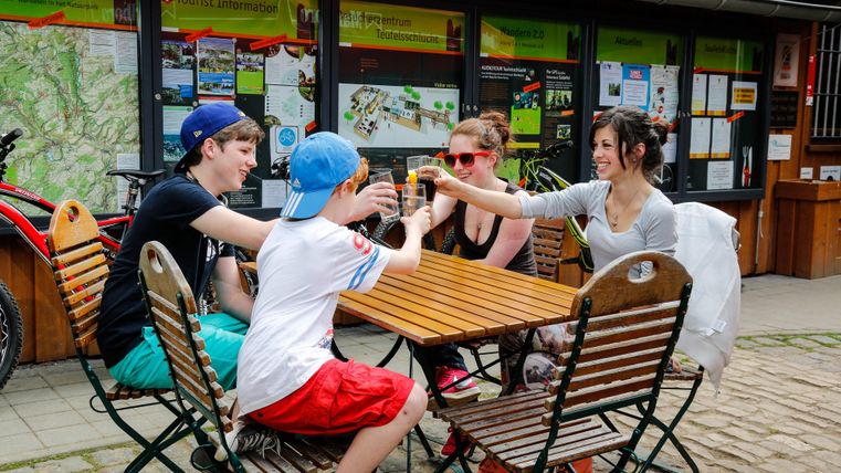 A group of four people sits at a table and raises glasses to toast. The background shows information about cycling in nature.