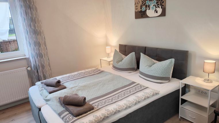 Ein gemütliches Schlafzimmer mit einem großen Bett und eleganten Kissen. Es gibt Fenster mit Vorhängen und Nachttische mit Lampen.