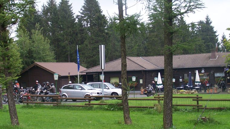 Ein gemütliches Gasthaus umgeben von Wald und Wiese. Vor dem Gebäude stehen Tische mit Sonnenschirmen und mehrere Fahrräder.