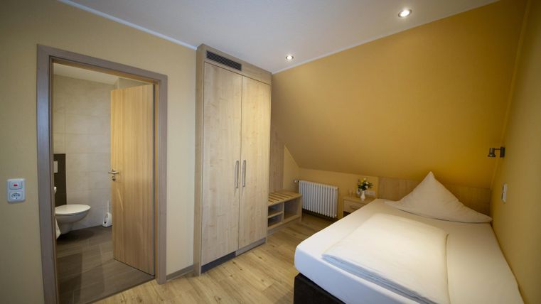 Ein einfaches Schlafzimmer mit einem Einzelbett und einem Kleiderschrank. Die Wände sind in warmen Gelbtönen gestrichen, und das Badezimmer ist durch eine Tür sichtbar.