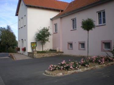 Ein charmantes Gebäude mit einem hellen Fassaden und einem orangefarbenen Dach. Vor dem Haus sind gepflegte Blumenbeete mit Rosen und kleinen Bäumen.