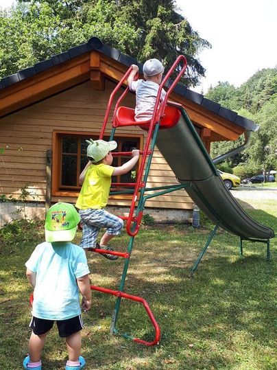 Zwei Kinder spielen auf einem Spielturm mit einer Rutsche im Freien. Im Hintergrund steht ein Holzhaus und die Sonne scheint.