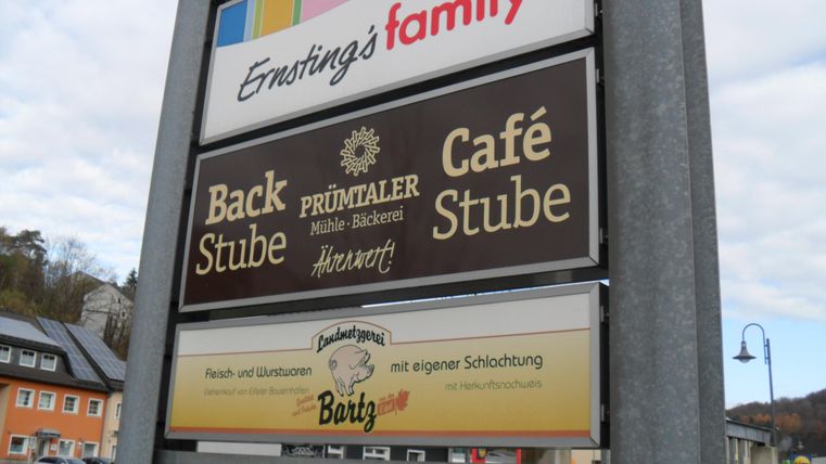 Das Bild zeigt ein Schild mit verschiedenen Geschäften. Zu sehen sind die Namen „Back Stube“, „Café Stube“ und „Prümtaaler Mühle-Bäckerei“.