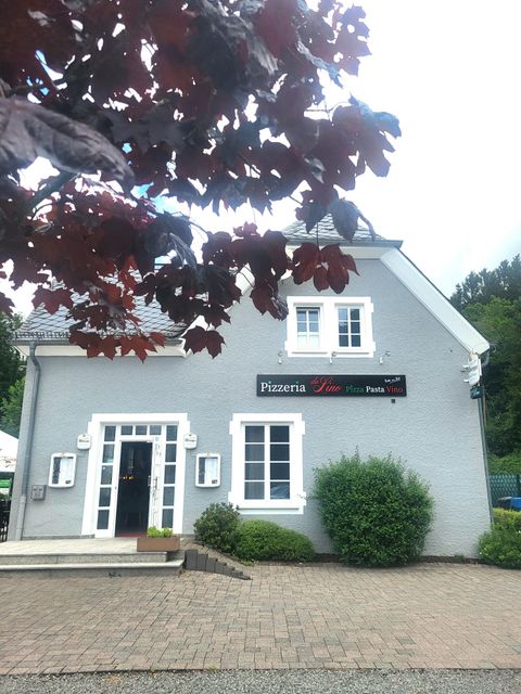 Ein graues Gebäude mit einem Schild, das "Pizzeria" anzeigt. Vor dem Eingang befinden sich Pflanzen und ein gepflasterter Weg.