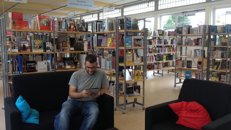 Ein Mann sitzt auf einem schwarzen Sofa in einer Bibliothek. Im Hintergrund sind Bücherregale mit vielen Büchern zu sehen.