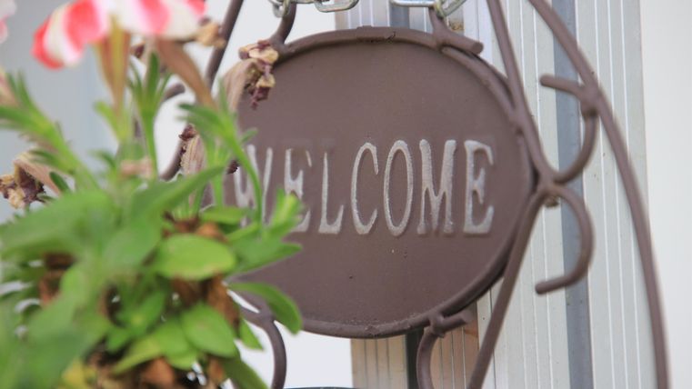 Ein Willkommensschild aus Metall hängt an einem Haken. Im Vordergrund sind Blumen zu sehen.