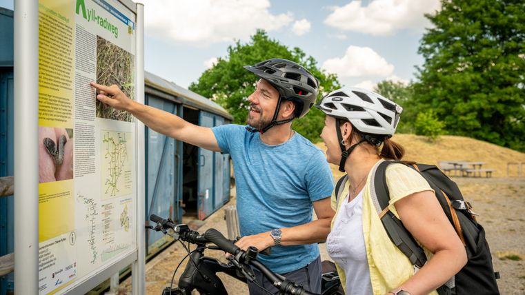 Zwei Radfahrer mit Helmen betrachten eine Infotafel am Kyll-Radweg.