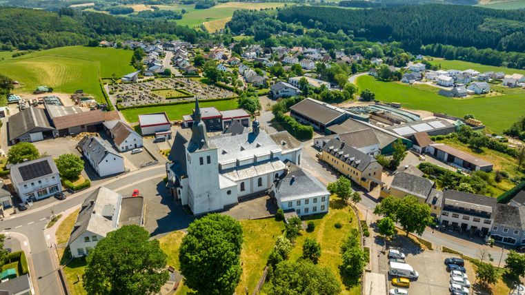 Eine malerische Landschaft mit einem weißen Kirchengebäude in der Mitte. Im Hintergrund sind grüne Felder und kleine Häuser zu sehen.