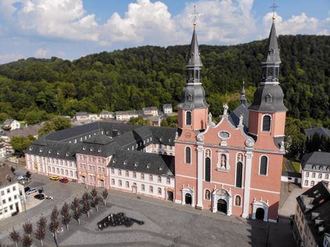 Eine beeindruckende Kirche mit zwei Türmen steht im Vordergrund. Im Hintergrund sind grüne Hügel und ein Platz mit Bäumen zu sehen.