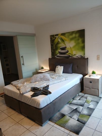 Ein ruhiges Schlafzimmer mit einem Bett und einer gemütlichen Bettdecke in grünen Tönen. An der Wand hängt ein Bild von Steinen und Pflanzen.