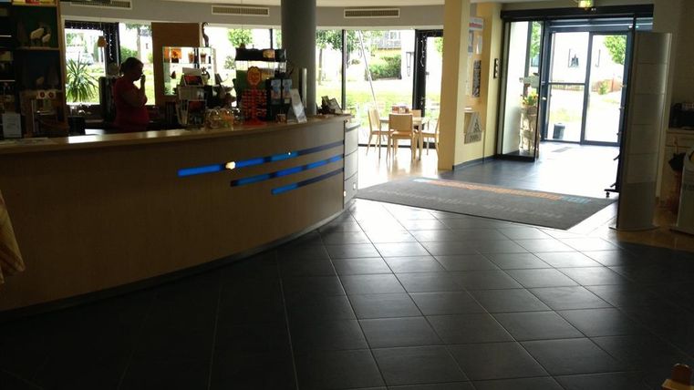 Ein Empfangsbereich mit einem großen, geschwungenen Empfangstresen. Im Hintergrund sind Tische und Stühle sowie Fenster, die auf einen begrünten Außenbereich blicken.