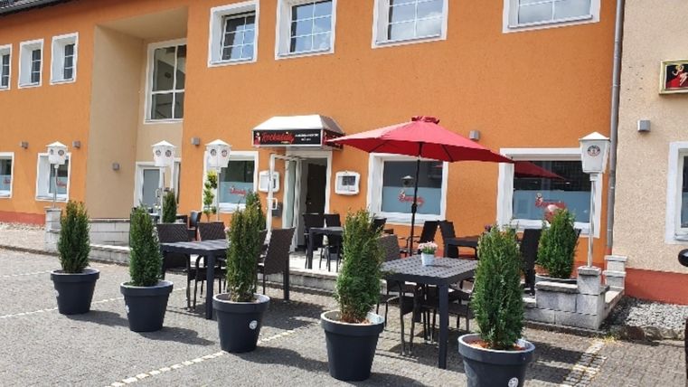 Ein modernes Gebäude mit orangefarbener Fassade und großen Fenstern. Draußen stehen Tische mit Stühlen und ein roter Sonnenschirm, umgeben von Pflanzen in schwarz-weißen Töpfen.