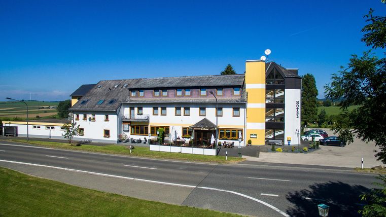 A modern hotel building on a street, surrounded by green spaces. The sky is clear and there are a few clouds visible.