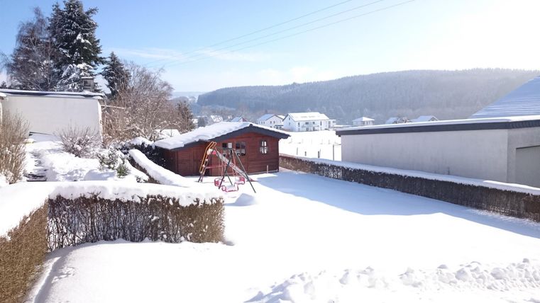 Eine schneebedeckte Landschaft mit einem roten Holzhaus im Hintergrund. Der Himmel ist klar und blau, und die Umgebung ist ruhig und winterlich.