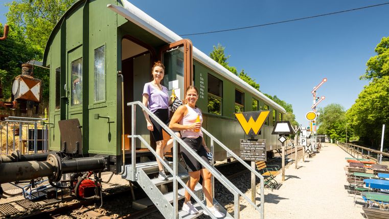 Zwei Frauen steigen aus einem alten Zugwagon und gehen die Treppe hinunter. Im Hintergrund sind Bahnschilder und eine grüne Umgebung zu sehen.