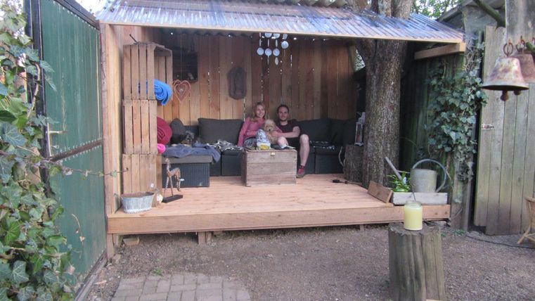 Eine gemütliche Holzterrasse mit zwei Personen auf einer Couch. Der Bereich ist von Pflanzen umgeben und hat ein rustikales Dach.