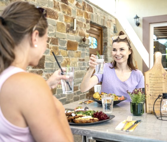 Zwei Frauen sitzen an einem Tisch im Freien, stoßen mit Getränken an und genießen eine Mahlzeit. Im Hintergrund eine Steinmauer., © Eifel Tourismus GmbH, Dominik Ketz