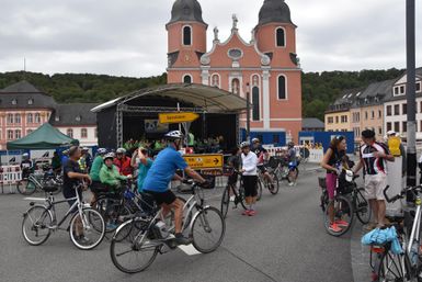 A group of cyclists gathers in front of a historic church. In the background, buildings and a stage can be seen.