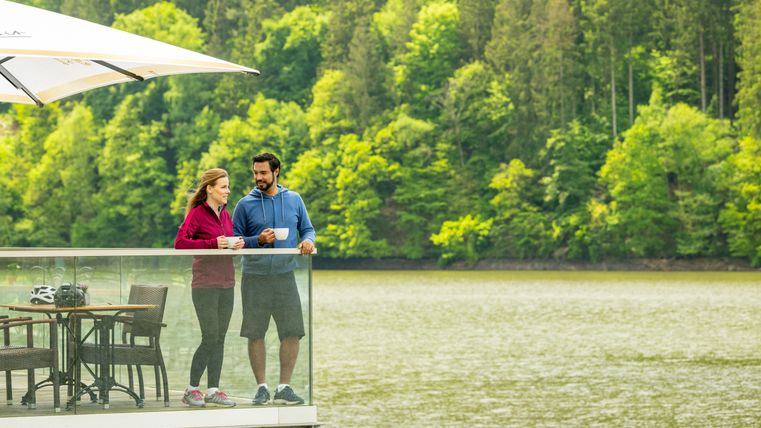 Eine Frau und ein Mann mit einer Tasse in der Hand stehen auf der Terrasse eines Cafés am Wasser