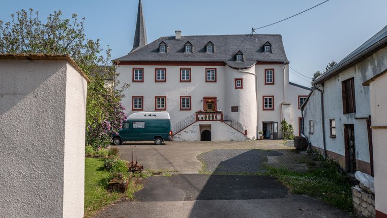 Ein historisches Gebäude mit einem spitzen Turm und einer großen Eingangsfront. Im Vordergrund stehen ein paar Fahrzeuge und grüne Landschaften.