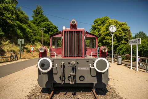 Eine rote Lokomotive steht an einem Bahngleis. Im Hintergrund sieht man Grünflächen und eine Uhr.