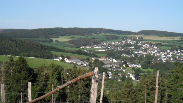 Eine malerische Landschaft mit sanften Hügeln und einem kleinen Dorf in der Mitte. Im Vordergrund sind Holzpfosten und Bäume sichtbar.