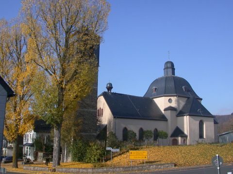 Eine Kirche mit einem runden Dach und einem hohen Turm, umgeben von Bäumen im Herbst. Die Farben der Blätter sind golden und die Himmel ist klar blau.