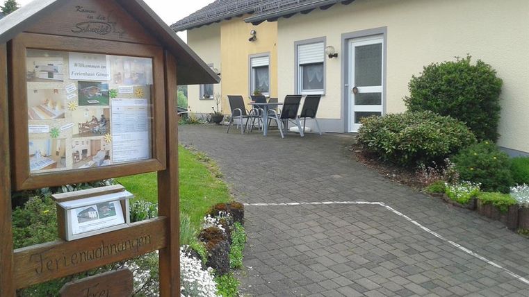 Ein gemütliches Ferienhaus mit einem gepflasterten Weg und einer einladenden Terrasse. Vor dem Haus steht ein Informationsschild über die Unterkunft.
