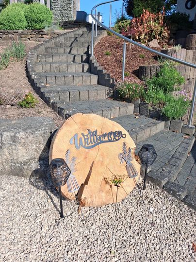Ein hölzernes Schild mit der Aufschrift "Willkommen" steht an einer Treppe. Im Hintergrund sind Pflanzen und Steine zu sehen.