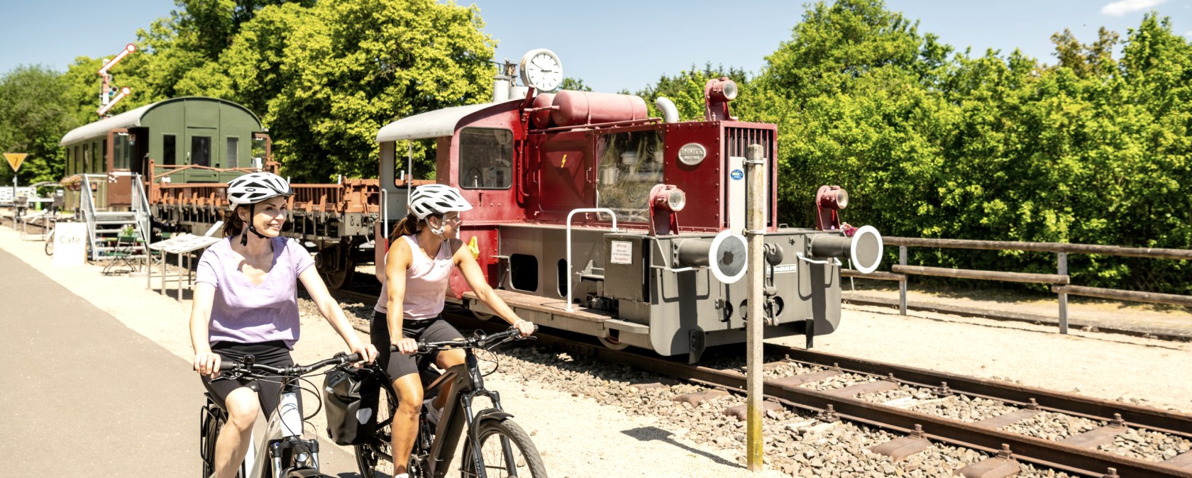 Zwei Radfahrerinnen auf einem Radweg neben einer alten Lokomotive im Eisenbahnmuseum Pronsfeld, umgeben von grünen Bäumen., © Eifel Tourismus GmbH, Dominik Ketz